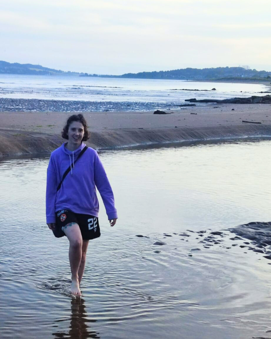 A person (Laura) smiling while walking through water on a sandy beach. Laura is wearing a purple jumper and black shorts.