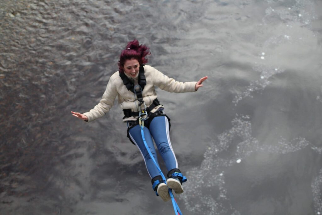 A person (Laura) doing a bungee jump above deep water. Laura is wearing a beige fleece and blue trousers.