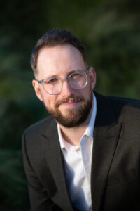 Jonathan Smith - A white man with brown hair, a beard and glasses is wearing a suit and smiling at the camera